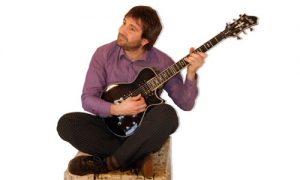 Daniel Brion est assis en tailleur et joue de la guitare électrique.